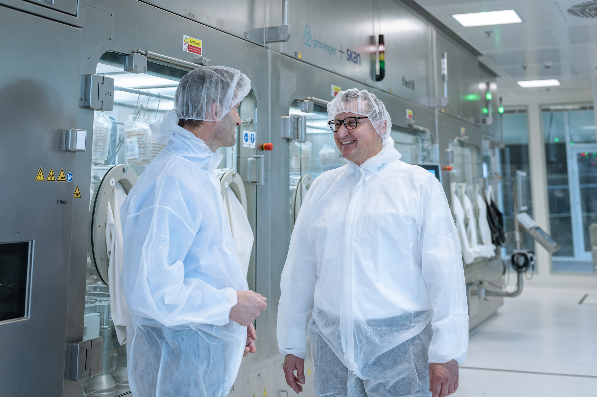 Two people in full cleanroom protective clothing stand facing each other and smiling while talking beside a row of stainless-steel laboratory isolator machines.