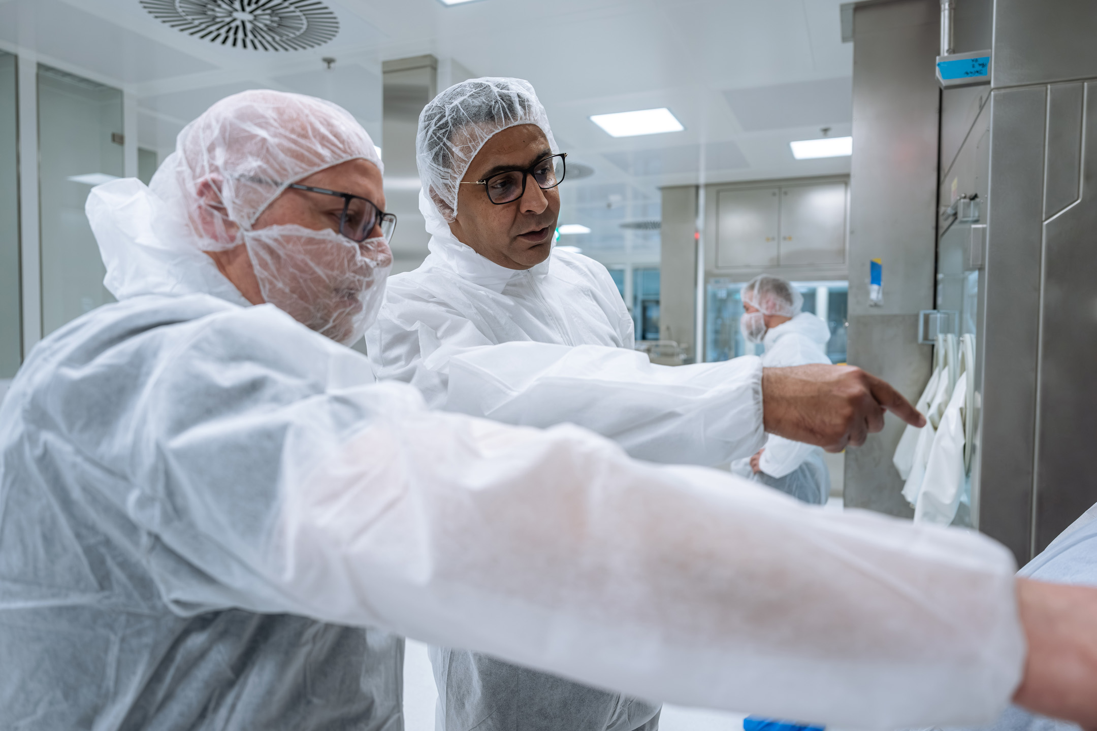Two people wearing cleanroom protective suits, hairnets, and glasses examine equipment closely, with one pointing toward a control or component inside a sterile laboratory environment.