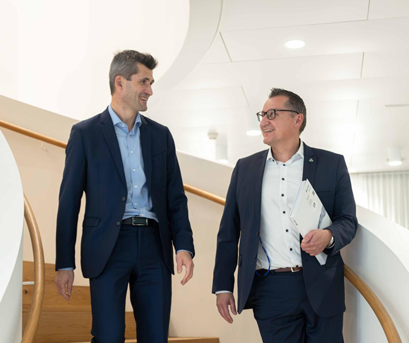 Two men in business suits walk down a modern staircase together, smiling and talking, while one holds a folder and a curved architectural wall frames the background.