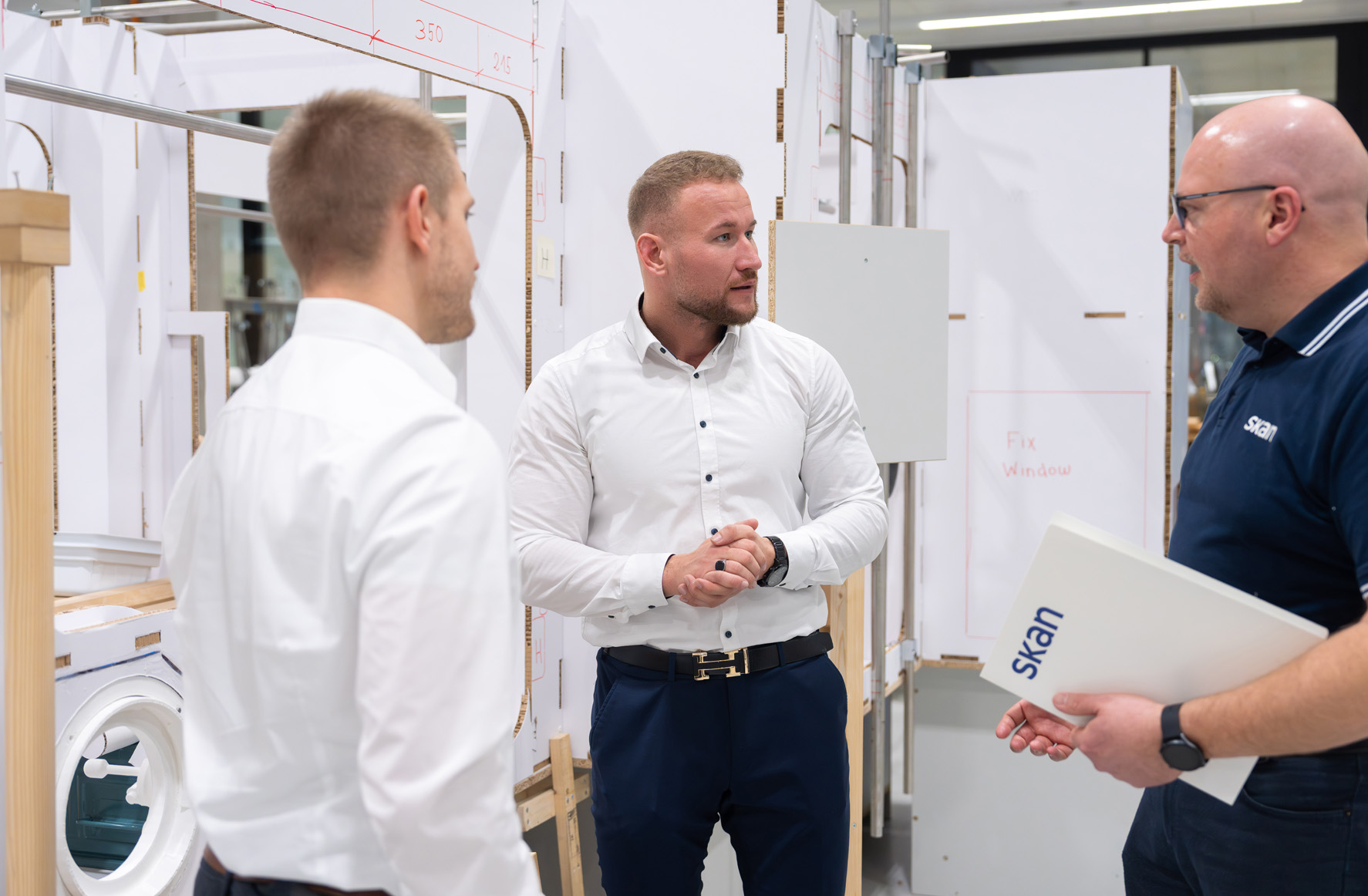 Three men stand together in a workshop or production area discussing something, while one holds a folder labeled “SKAN” and equipment or structural components are visible in the background.